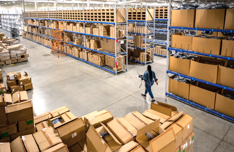 Worker walking through a warehouse with shelving units stacked with cardboard boxes for storage and distribution.