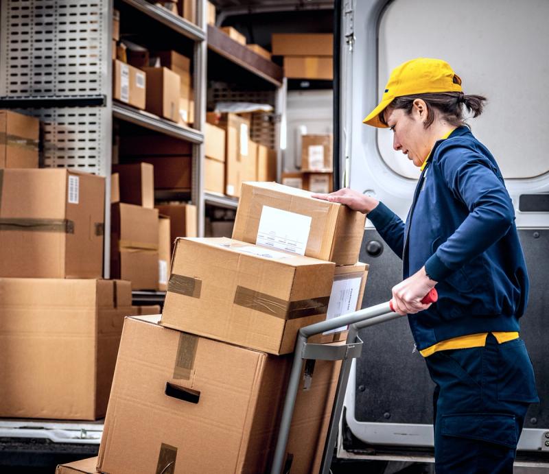 Delivery courier in a yellow cap loading parcels from a hand truck into a van