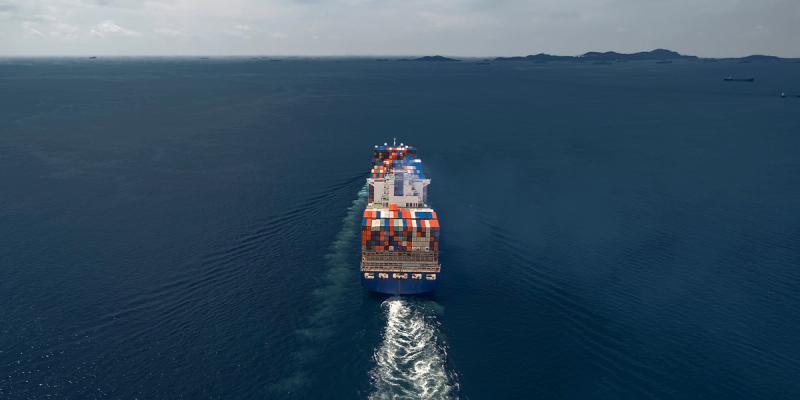 Aerial view of a loaded container ship sailing away on calm open sea toward distant islands