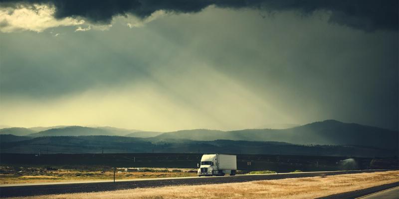 White semi-truck on a rural highway under a dramatic stormy sky with sunbeams over distant hills.