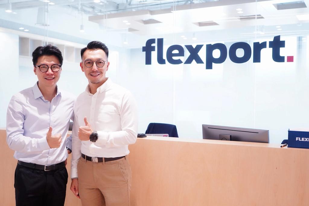 Two men give thumbs up in a Flexport office in front of a wall with the flexport logo