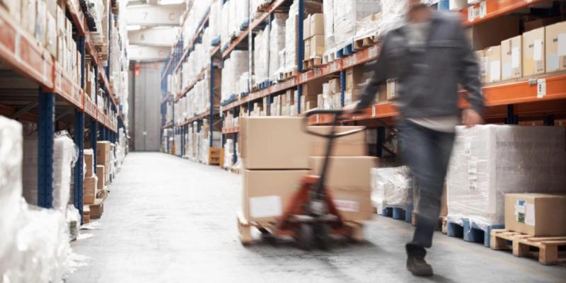 Worker pushing a pallet jack of boxes down an aisle between tall stocked warehouse racks, motion blur