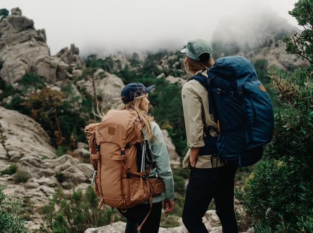 Two hikers with large Fjällräven backpacks pause on a rocky mountain trail with mist over distant peaks