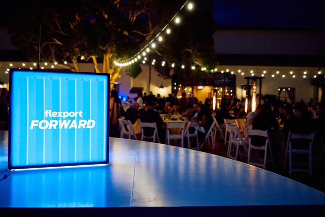 Illuminated 'Flexport FORWARD' sign on a table at an outdoor evening event with string lights and diners