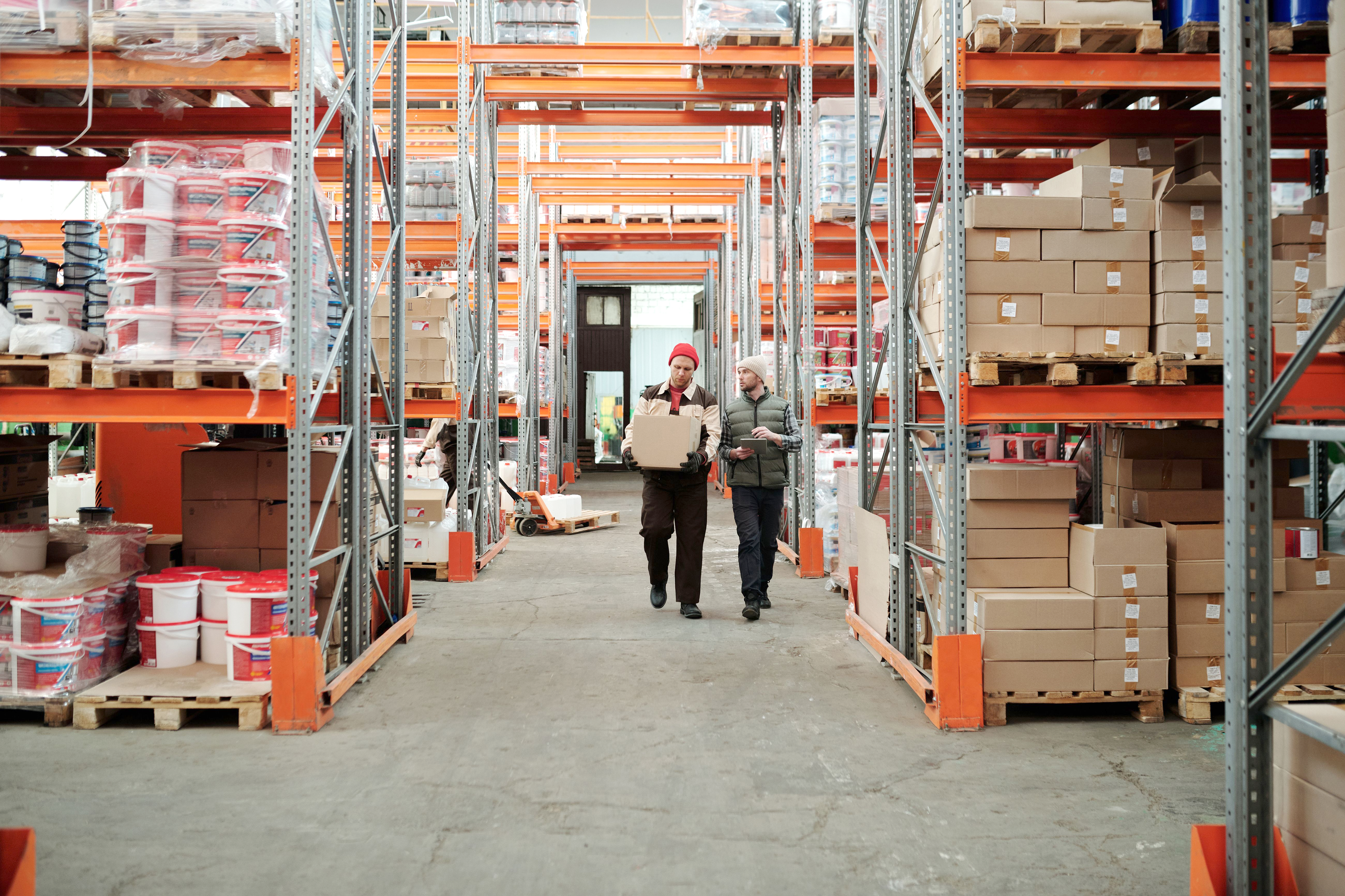 Two warehouse workers walking through a storage facility carrying boxes between shelving units.
