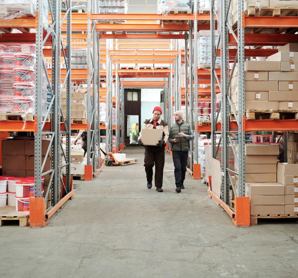 Two warehouse workers walking through a storage facility carrying boxes between shelving units.