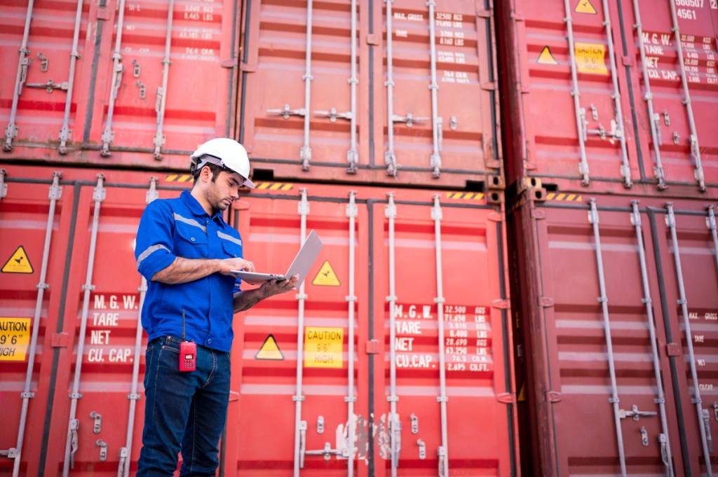 Worker in blue coveralls and white hard hat using a laptop in front of stacked red shipping containers