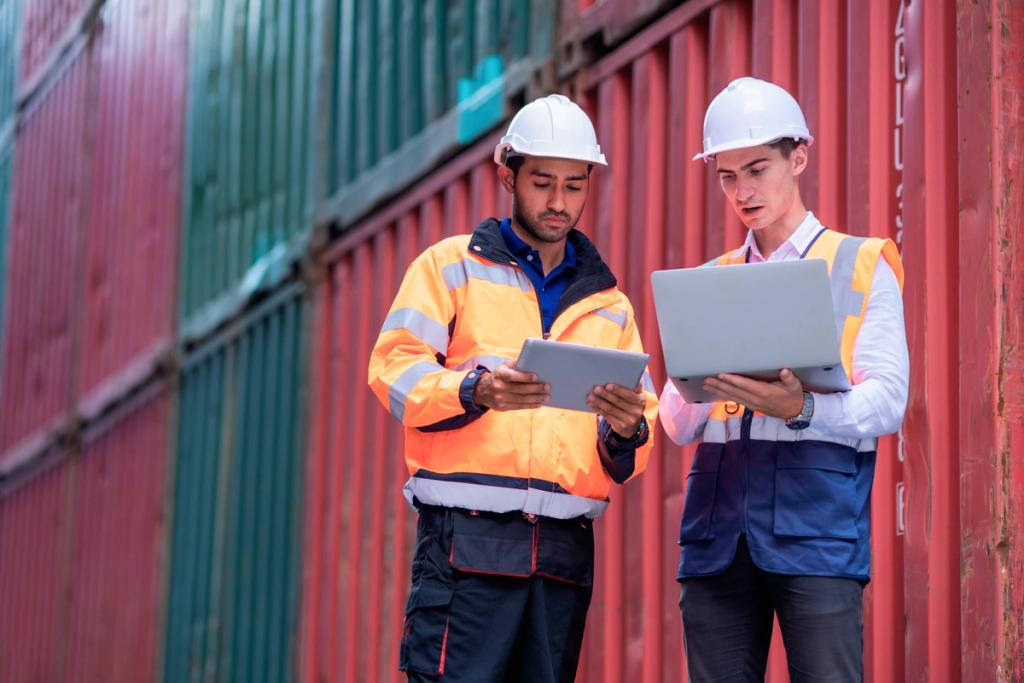Two workers in hard hats and hi-vis vests reviewing a tablet and laptop in front of red shipping containers