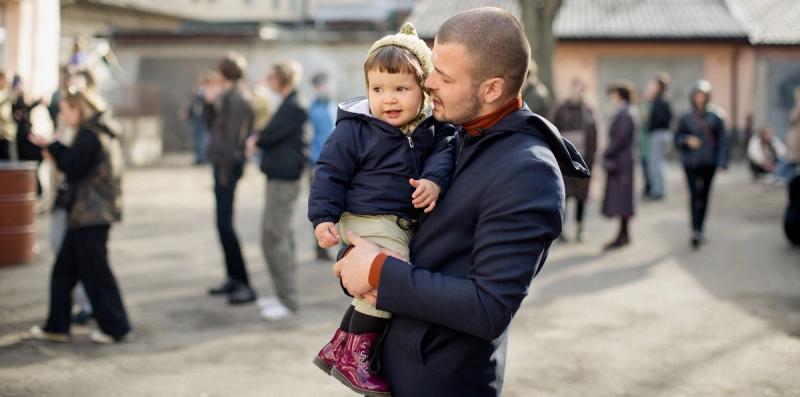 A man holds a small smiling child wearing a knit hat among a crowd of people gathered outdoors