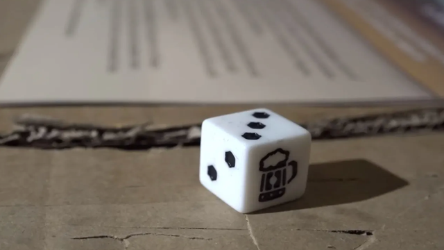 White six-sided die with a beer-mug icon on one face, resting on a kraft cardboard surface with printed text behind it