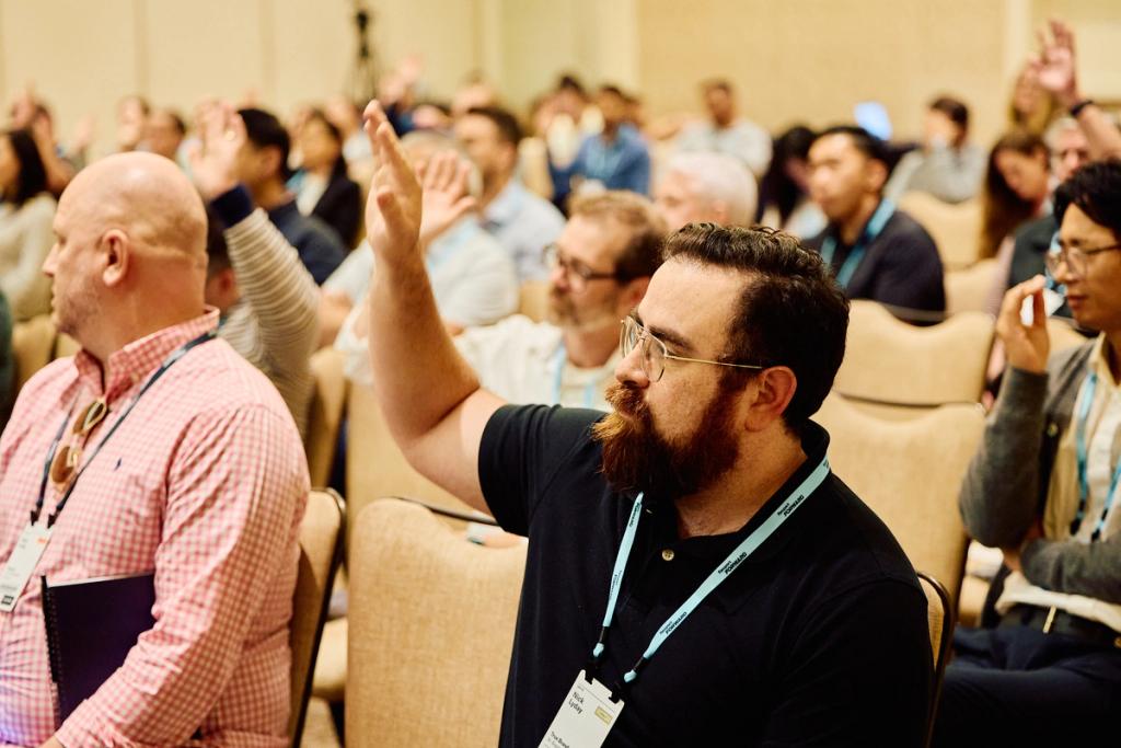 Bearded conference attendee raises his hand among seated audience members wearing lanyards in a ballroom.