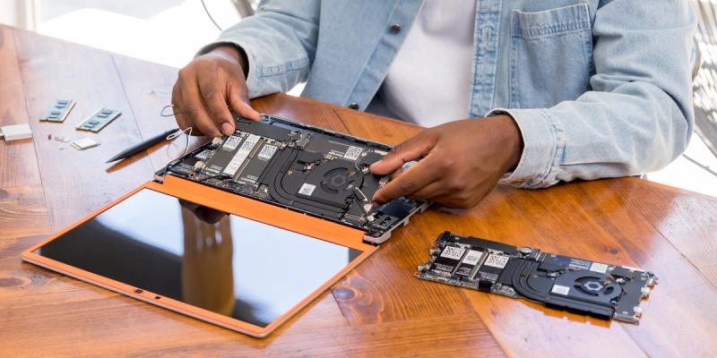 Person in denim repairs an open Framework laptop, with internal components and a spare mainboard on a wooden table
