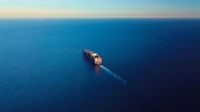 Aerial view of a container ship laden with cargo crossing deep blue open ocean, leaving a wake behind it