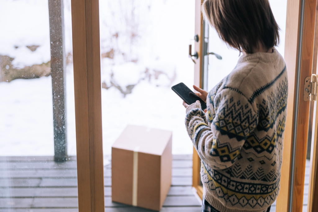 Person in patterned sweater checks a phone at an open door with a parcel on a snowy porch