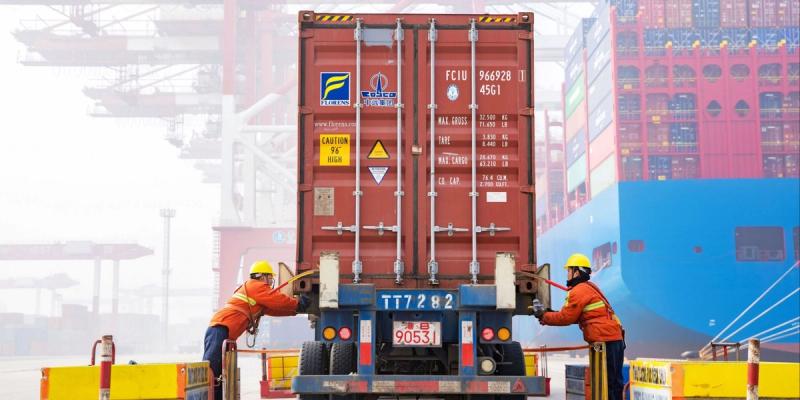Two dockworkers in orange vests and hard hats securing a red Florens shipping container on a truck at a foggy port