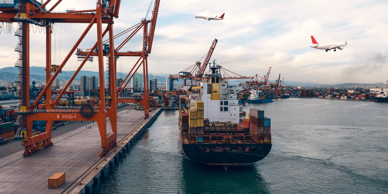 Container ship leaving a port with gantry cranes as two airplanes fly overhead