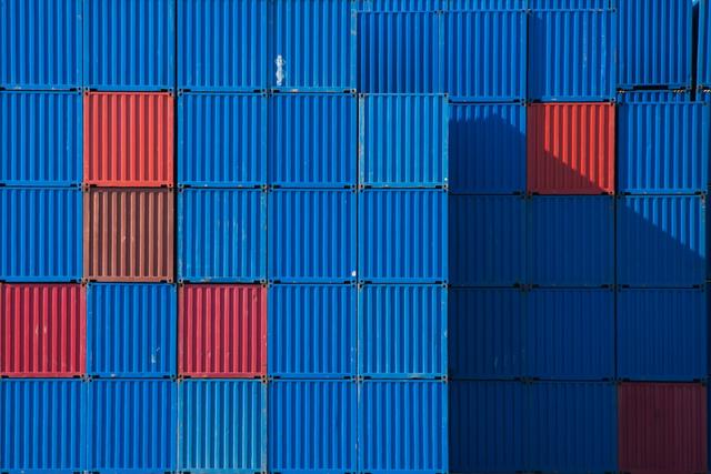 Stacked shipping containers filling the frame, mostly blue with red and maroon accents, diagonal shadow across the right side