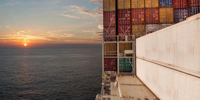 View from a container ship's deck of stacked containers and white hull walls as the sun sets over the open ocean.