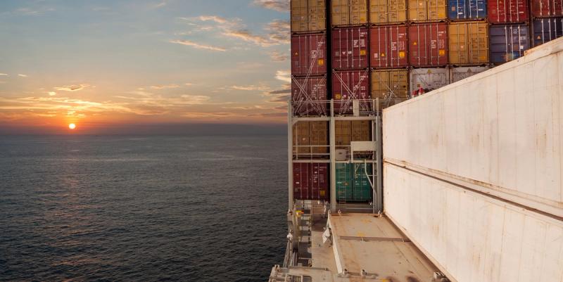 View from a container ship's deck of stacked containers and white hull walls as the sun sets over the open ocean.