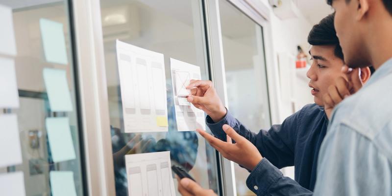 Two men review printed UI wireframes taped to a glass wall, one pointing while holding a phone
