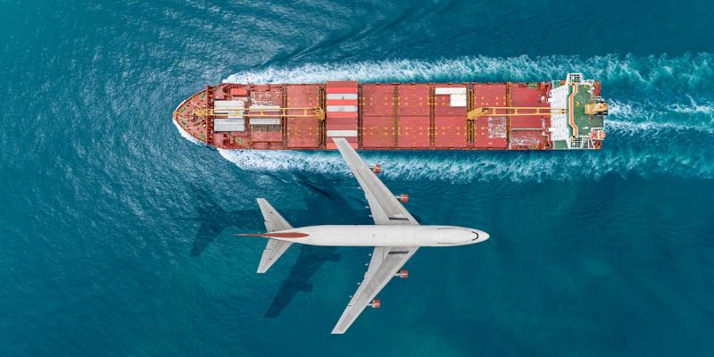 Aerial view of a passenger jet flying directly over a red container ship on teal ocean water