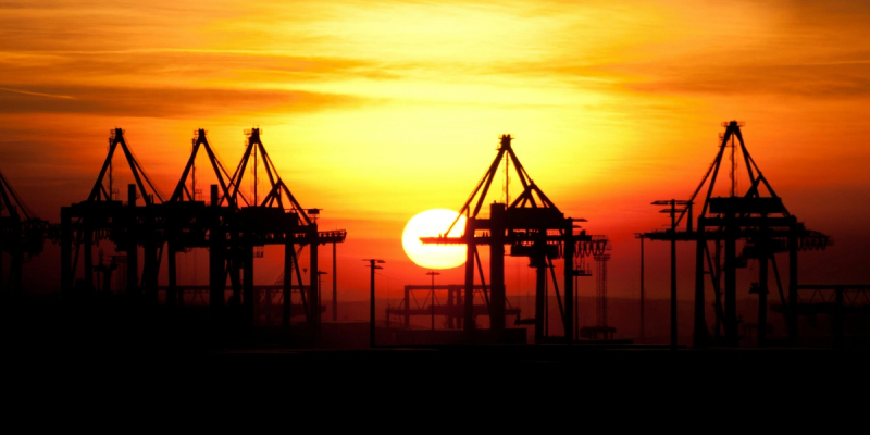Silhouettes of port gantry cranes against a vivid orange sunset sky with the sun on the horizon