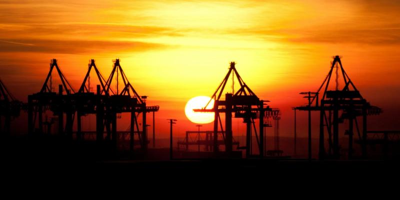 Silhouettes of port gantry cranes against a vivid orange sunset sky with the sun on the horizon