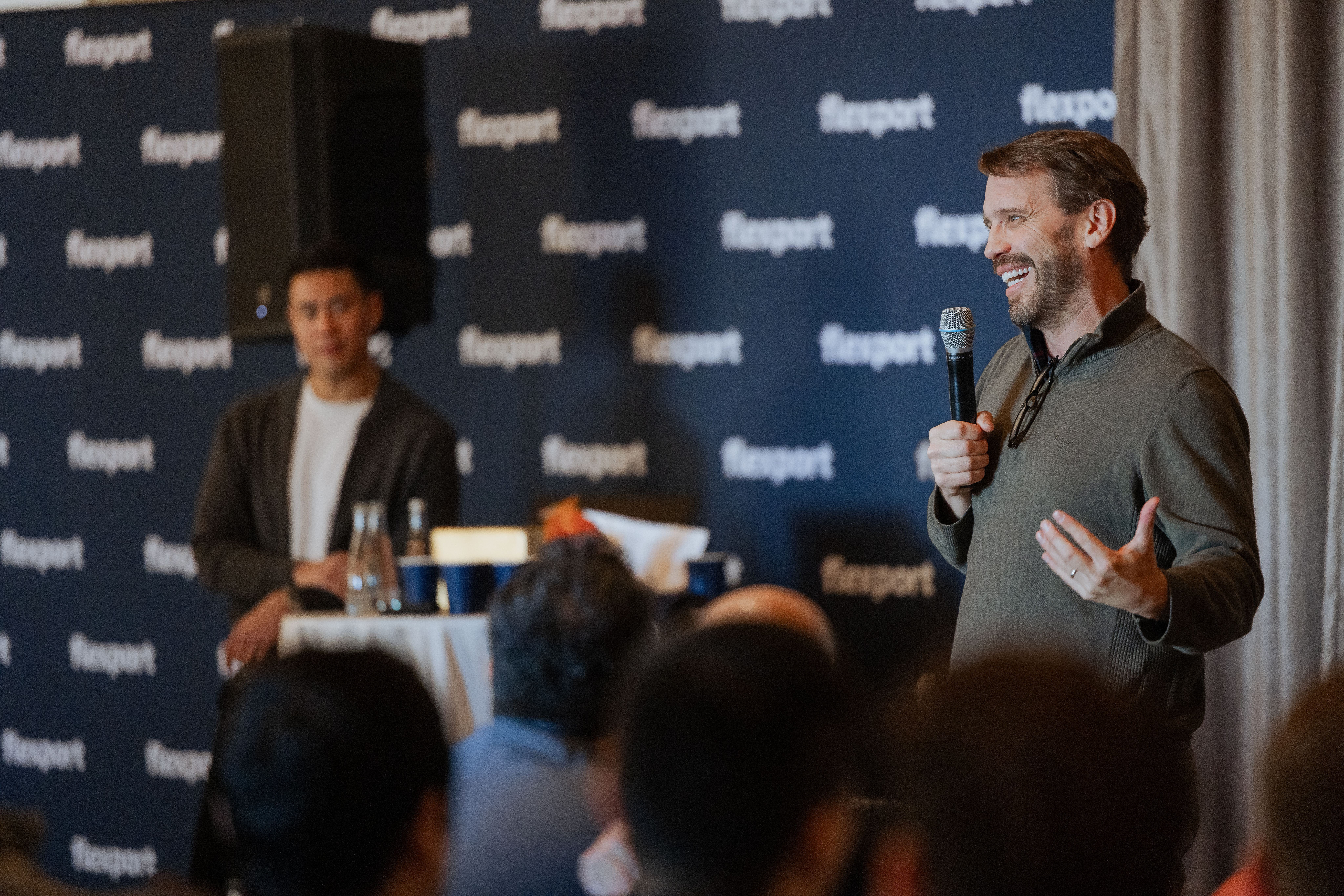A man speaks into a microphone at a panel discussion while audience members listen.
