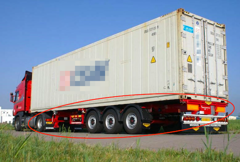 A red semi-truck hauling a beige ocean shipping container loaded on a chassis trailer along a road, the chassis frame outlined in red.