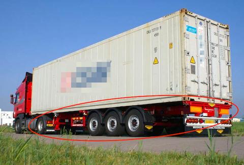 A red semi-truck hauling a beige ocean shipping container loaded on a chassis trailer along a road, the chassis frame outlined in red.