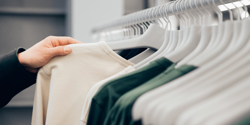Hand examines a cream t-shirt on a rack of white and green apparel on hangers