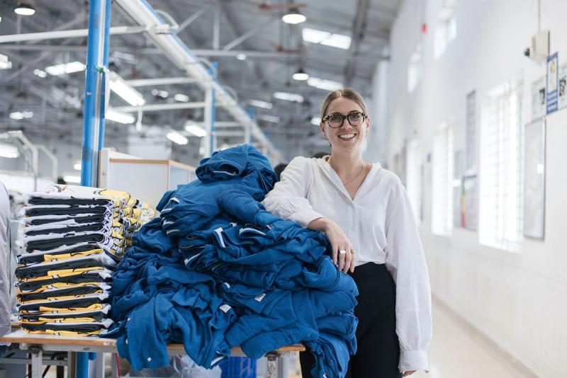 Smiling woman in a white blouse stands by a stack of folded blue garments in a bright apparel factory