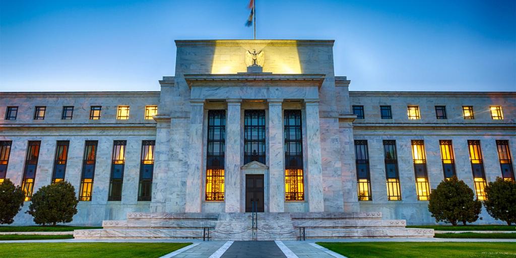 Front facade of the marble US Federal Reserve building lit at dusk, with an eagle relief above the central columns.
