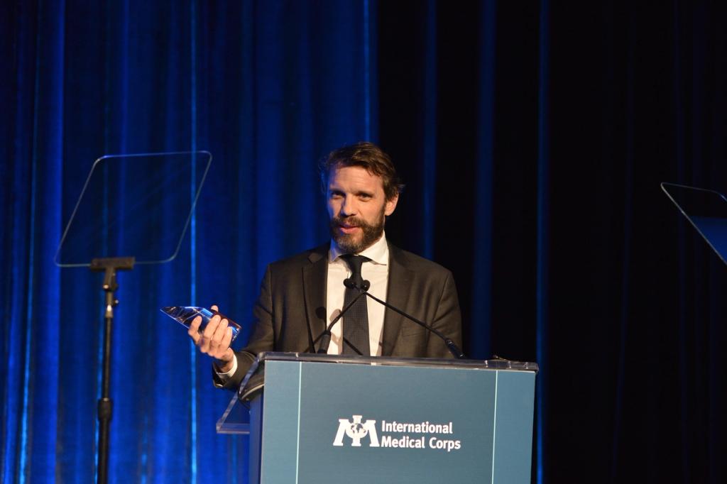 Bearded man in a suit holds an award at a lectern marked International Medical Corps