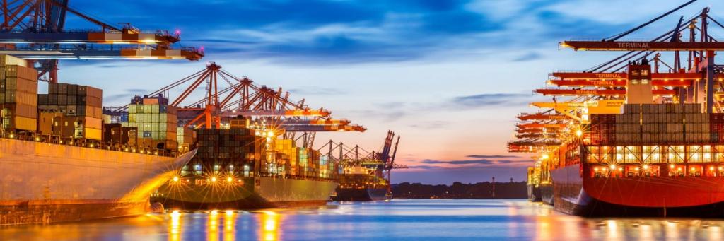 Container ships loading at a port terminal at dusk under blue sky with illuminated cranes reflecting on water