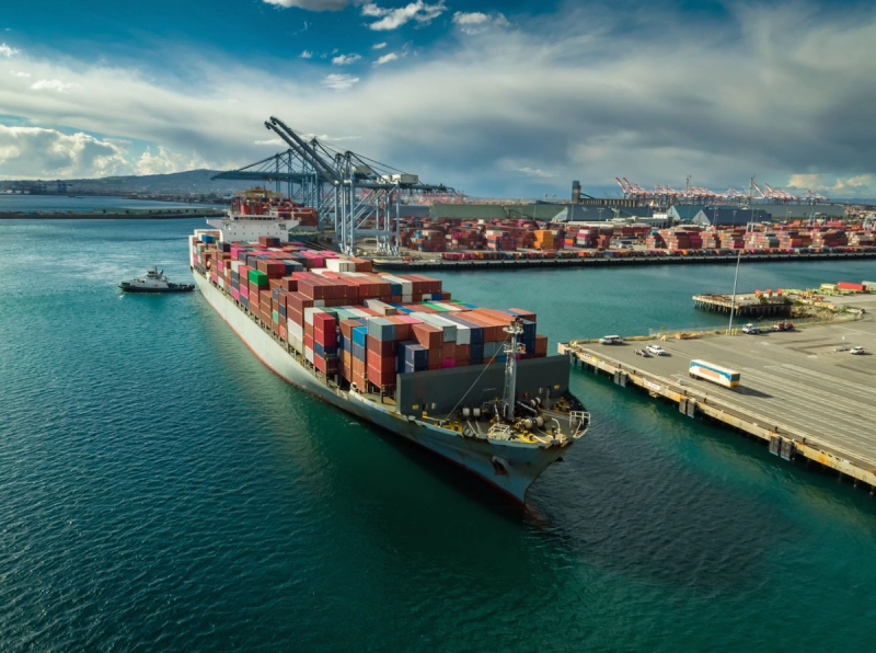 Aerial view of a container ship loaded with stacked containers docking beside port cranes and a tugboat