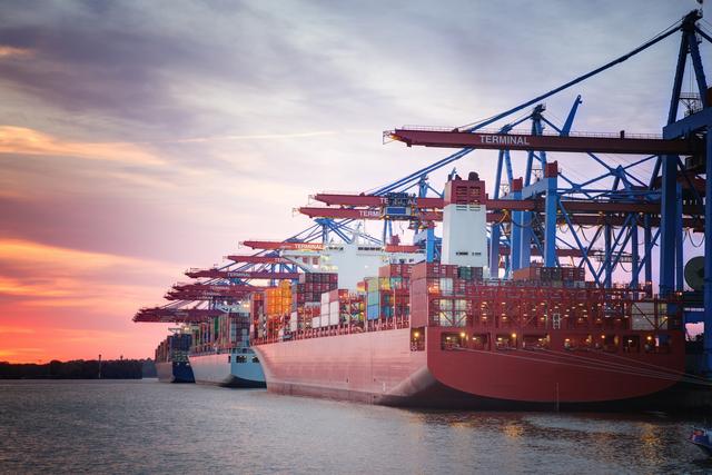 Container ships berthed at a port terminal under cranes labeled TERMINAL at sunset with orange sky reflected in the water