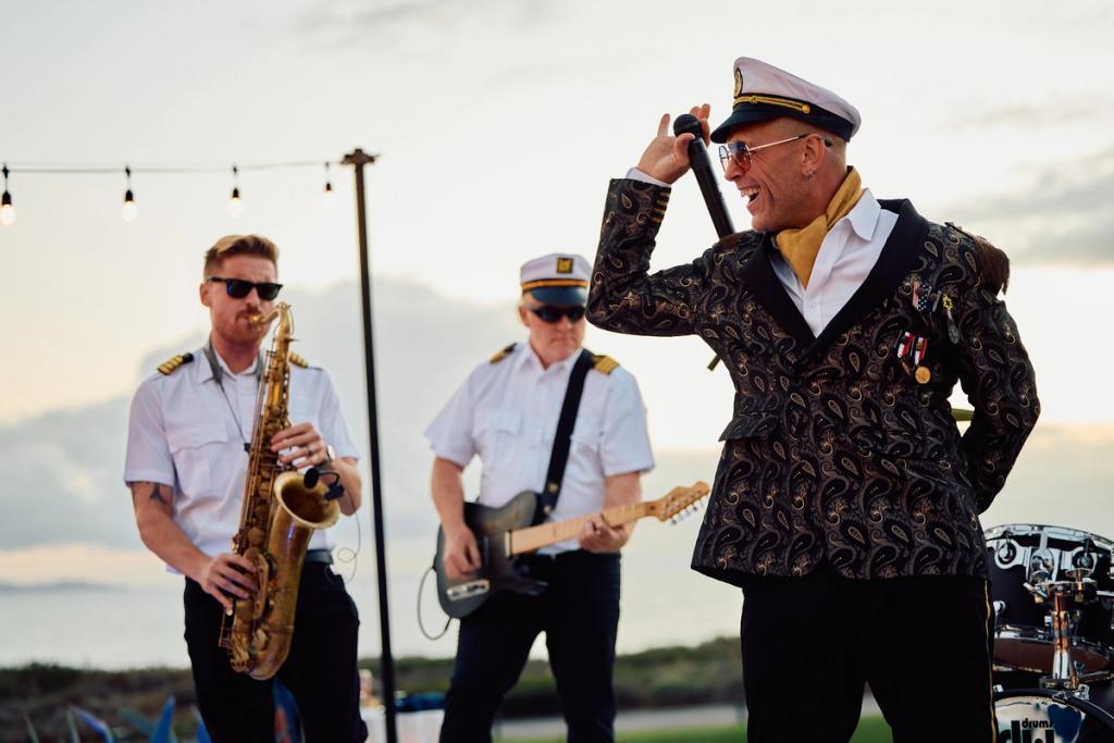 Live band in naval-style captain caps performs at sunset, vocalist in a paisley jacket singing into a mic