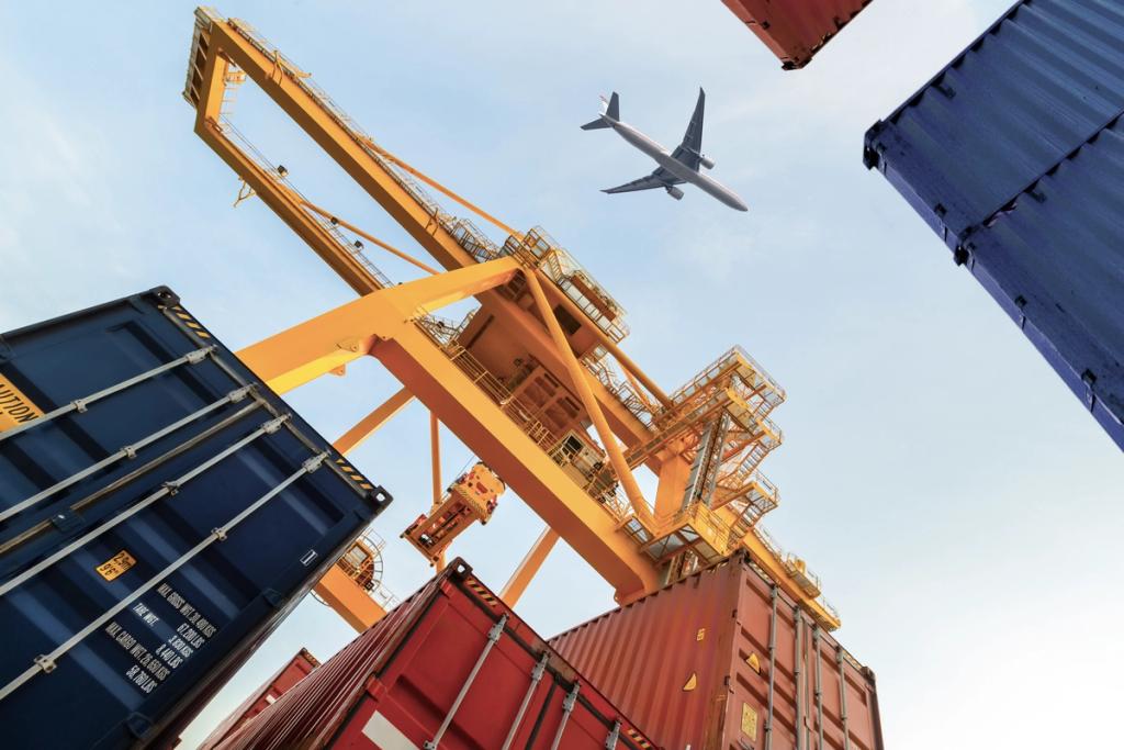 Upward view of a yellow port gantry crane between stacked shipping containers with an airplane passing overhead in a clear sky