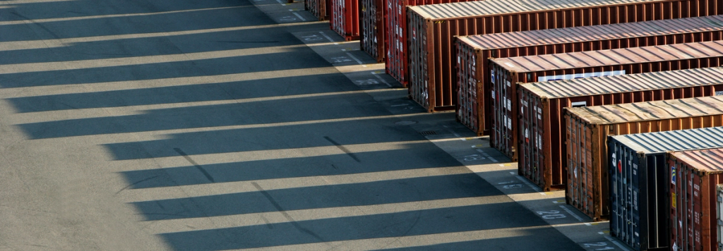 Rows of stacked shipping containers at a port terminal with long shadows across the asphalt
