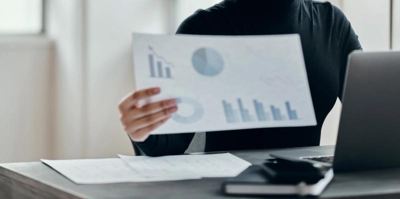 Person in dark clothing holding a printed page of bar charts and pie charts at a desk with a laptop