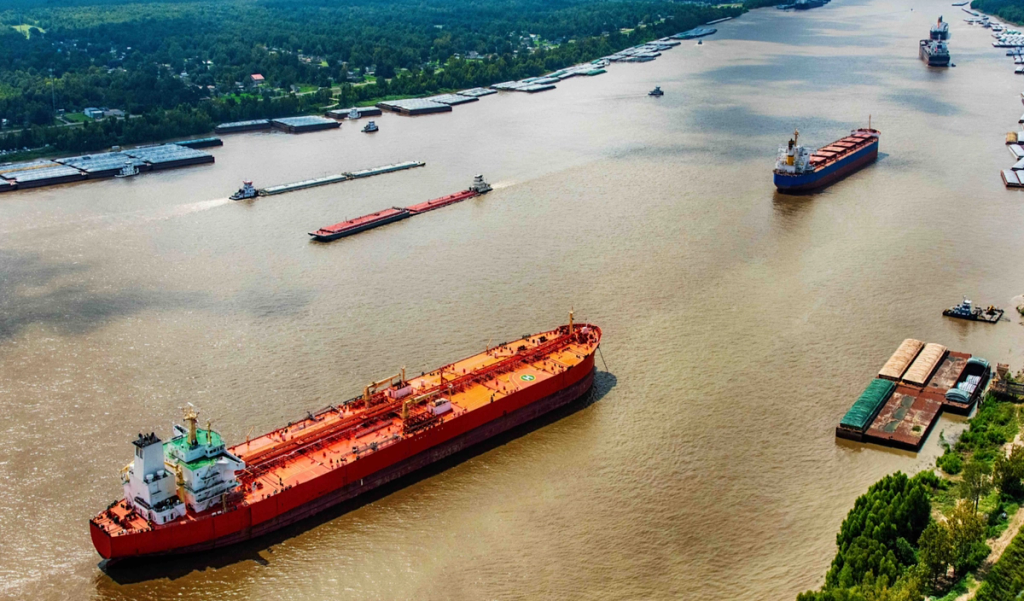 Aerial view of barges and an orange oil tanker navigating a wide green river lined with forest