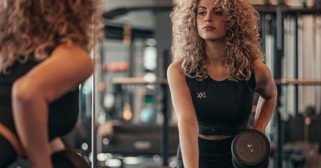 Woman with curly blonde hair in XXL workout top lifting a dumbbell in front of a gym mirror