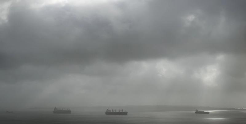 Three cargo ships at anchor on calm water beneath a heavy overcast grey sky