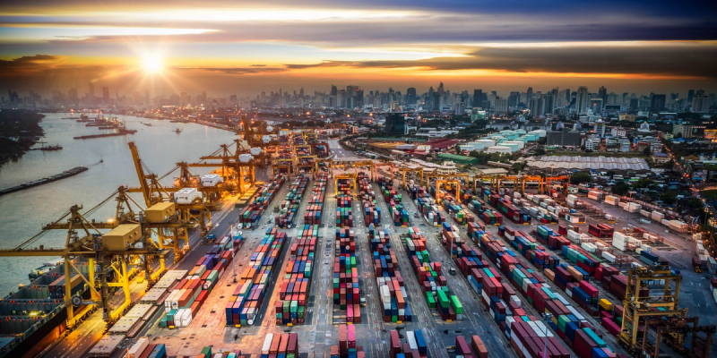 Aerial view of a sprawling container port and city skyline at sunset with cranes and rows of containers