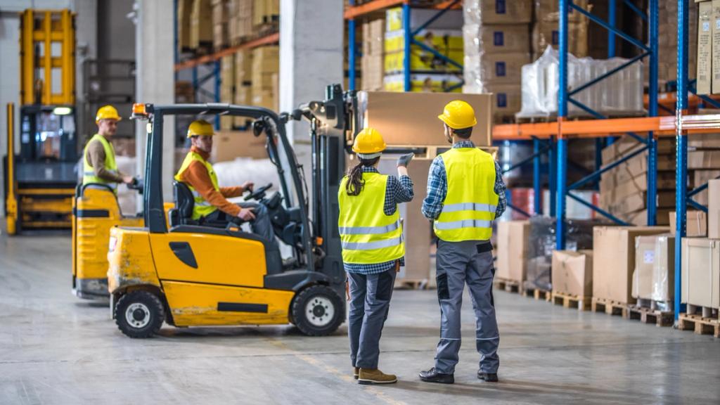 Warehouse interior with a forklift carrying a box and two workers in high-vis vests and hard hats among racked pallets