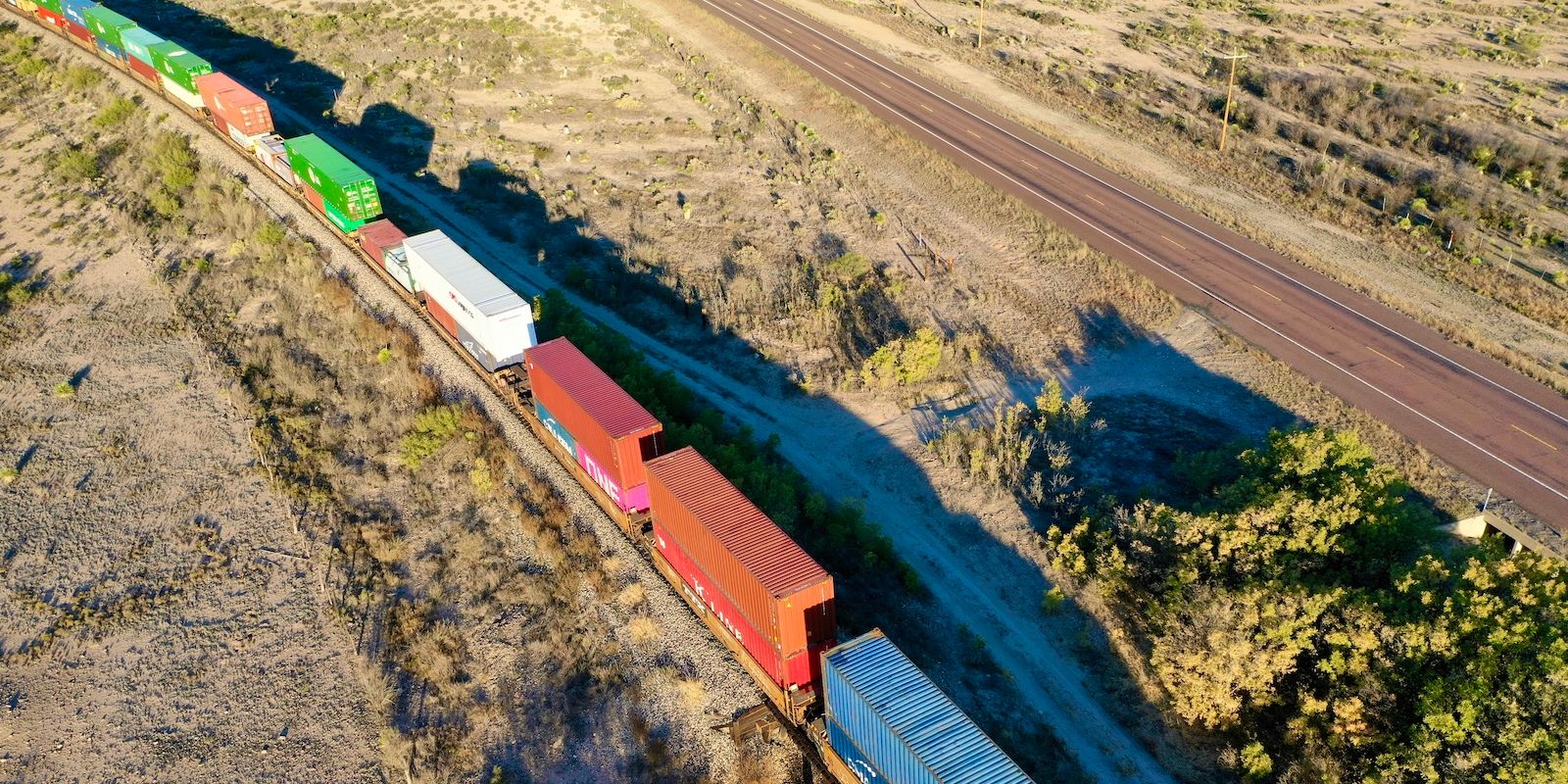 Aerial view of a freight train of stacked intermodal containers crossing arid desert beside a highway