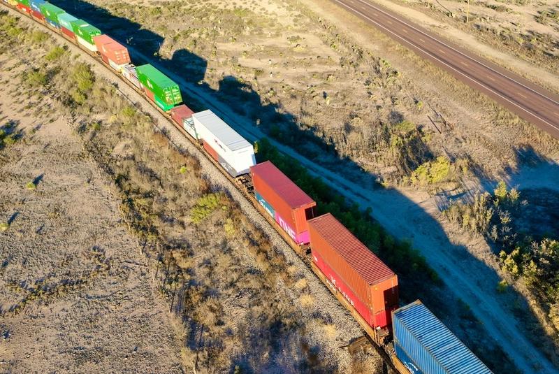 Aerial view of a freight train of stacked intermodal containers crossing arid desert beside a highway