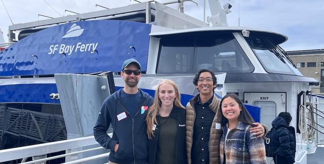 Four people smiling pose in front of a blue SF Bay Ferry catamaran at a dock.