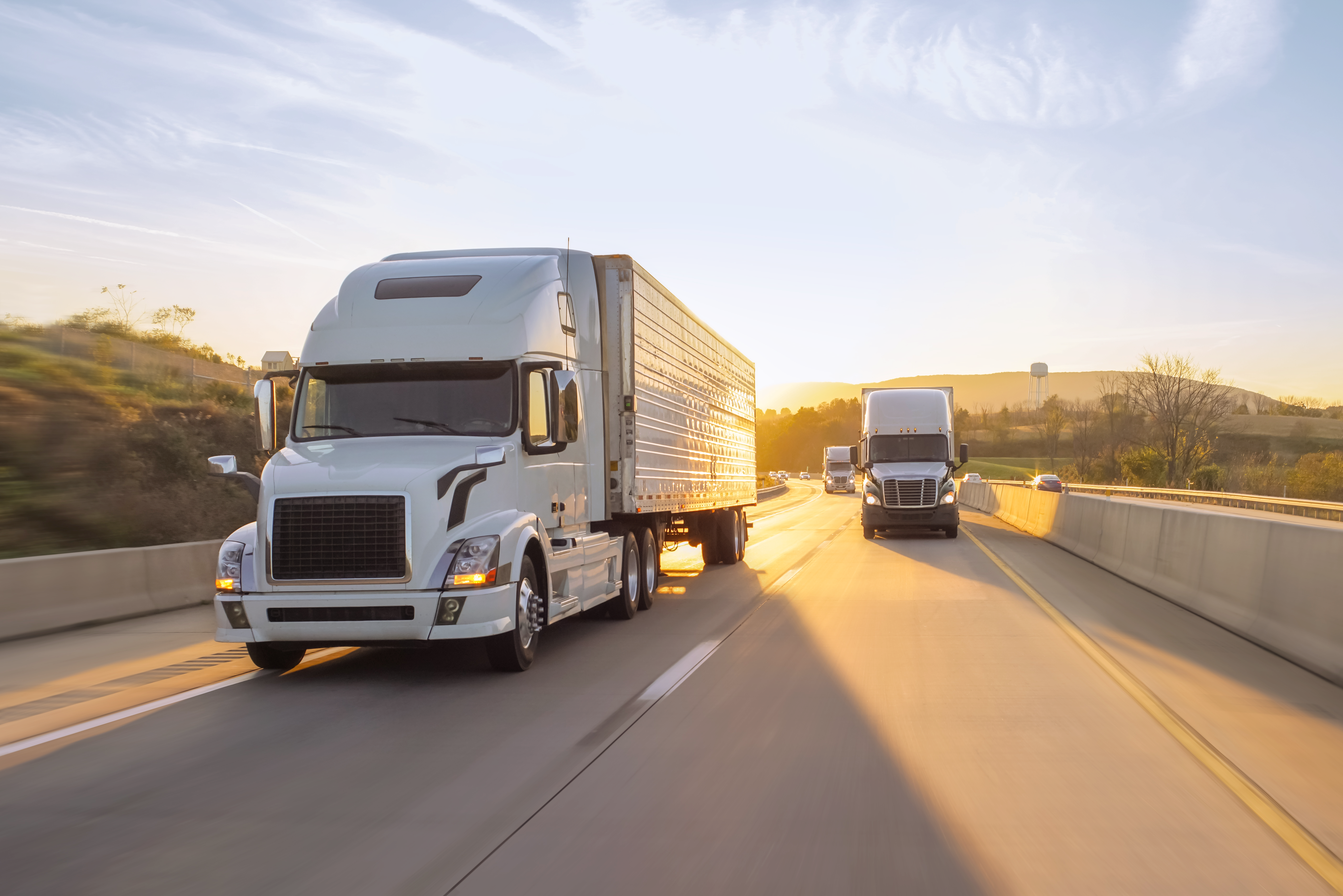 White semi trucks hauling trailers down a highway into a low sunset